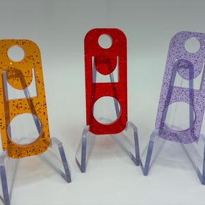 Op de afbeelding: Drie rechthoekige acryllabels met een glitterafwerking, elk voorzien van een rond gat aan de bovenkant en een grotere ronde uitsparing aan de onderkant. De oranje, rode en paarse labels staan rechtop op doorzichtige kunststof standaards.