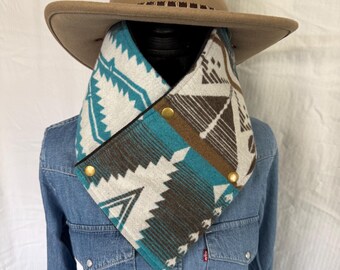 Könnte beinhalten: Ein beigefarbener Cowboyhut sitzt auf einer Schaufensterpuppe, die ein Jeanshemd und einen gemusterten Schal trägt. Der Schal hat ein geometrisches Design in Türkis, Braun und Weiß und ist mit goldfarbenen Knöpfen befestigt. Der Hut hat ein braunes Lederband.