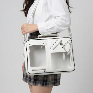 Puede incluir: Bolso mensajero blanco con paneles frontales transparentes, herrajes plateados y un detalle de cadena. El bolso tiene un asa superior y una correa para el hombro. El bolso lo lleva una persona.