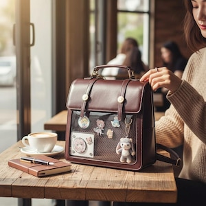 Op de afbeelding: Een bruine leren rugzak met een doorzichtige voorzak met kleurrijke spelden en een klein knuffeldier. De tas heeft een handvat aan de bovenkant, verstelbare bandjes en goudkleurige hardware. Een notitieboek en een kopje koffie staan op een houten tafel.