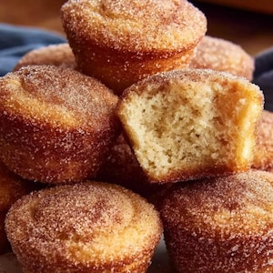 May include: A stack of golden-brown muffins, coated in a generous layer of cinnamon sugar. One muffin has a bite taken out, revealing the soft, airy interior. The baked goods are piled on top of each other.