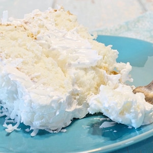 May include: A slice of coconut cake with white frosting and shredded coconut on a blue plate. The cake has a fluffy texture and is topped with a generous amount of frosting. A fork is visible on the right side of the plate.