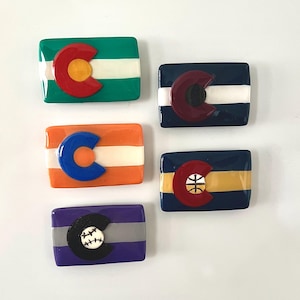 Könnte beinhalten: Rechteckige Magnete mit dem Design der Colorado-Flagge. Jeder Magnet zeigt das "C"-Logo des Staates mit verschiedenen Sportmotiven, darunter Baseball, Basketball und einfarbige Versionen. Die Magnete sind in Grün, Orange, Marineblau und Lila gehalten.
