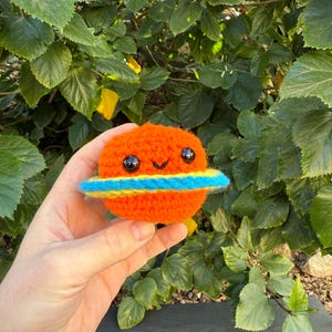 May include: An orange crocheted planet with a blue and yellow ring. The planet has two black eyes and a smiling mouth. The handmade toy is held in front of a background of green leaves.