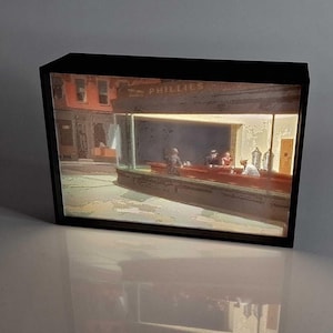 Puede incluir: Una caja de luz rectangular negra que muestra una pintura de una escena de restaurante. La pintura presenta una barra roja, figuras y la palabra "PHILLIES" en el toldo. La escena está iluminada, creando un brillo cálido.