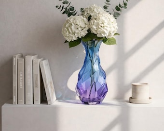 May include: A faceted glass vase with a gradient of blue to purple holds white hydrangeas and eucalyptus. The vase sits on a white shelf with books and a small ceramic cup.