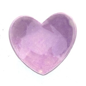 May include: A heart-shaped, lavender-colored dish. The dish has a smooth, glossy surface and is set against a white background. The heart shape is clearly defined, with rounded edges and a shallow interior.