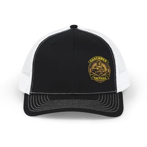 May include: Black and white trucker cap with a curved peak and mesh back. The front panel is black with a gold emblem reading "Oakember Tactical". The emblem depicts a figure holding a weapon.