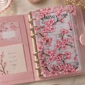May include: A light pink six-ring binder featuring a clear plastic cover with a pink cherry blossom print. Inside, a sinking funds tracker sheet is visible, along with a card and a note that reads, take time to make your soul happy.