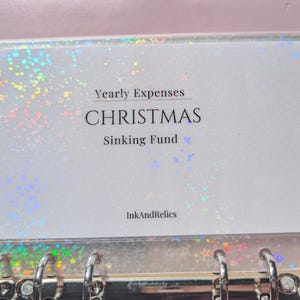 Może przedstawiać: Różowy segregator z przezroczystą kieszenią, w której znajduje się biała wkładka. Wkładka zawiera napis "Yearly Expenses CHRISTMAS Sinking Fund" czarnym tekstem. Przezroczysta kieszeń ma holograficzny, błyszczący wzór. Marka "InkAndRelics" znajduje się na dole.