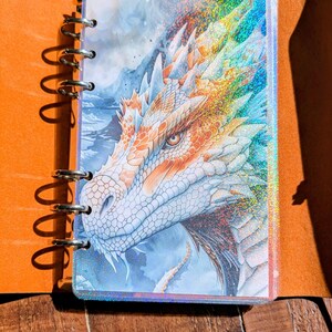 Peut inclure: Un journal sur le thème de la fantaisie avec une illustration de dragon. Le journal présente une tête de dragon détaillée avec des teintes orange, blanches et bleues. La couverture a un effet holographique. Le journal est relié dans une couverture en cuir marron avec un charme de feuille.
