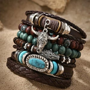 Könnte beinhalten: Ein Stapel brauner Lederarmbänder mit silbernen Akzenten, Türkissteinen und Holzperlen. Ein Armband hat einen silbernen Bullenschädel-Anhänger. Die Armbänder sind auf einer sandigen Oberfläche gestapelt.