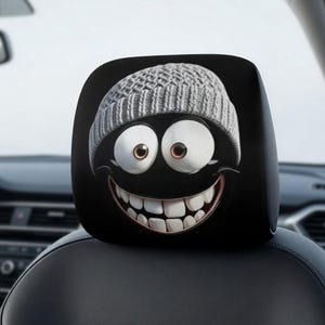 以下が含まれることがあります： 黒い車のヘッドレストカバー。グレーのニット帽、大きな白い目、大きく笑った口の漫画の顔が描かれています。ヘッドレストは黒い車のシートに取り付けられています。