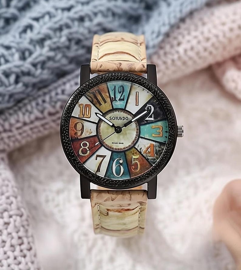 May include: A round wristwatch with a black bezel and a colorful face featuring large numerals. The watch has a light-colored band with a textured pattern. The watch face displays the brand name "SOTUKDO".