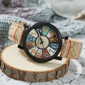 May include: A round wristwatch with a black case and a colorful face featuring the text "SONSDO". The watch has a cork band with colorful stripes and is displayed on a wooden slice.