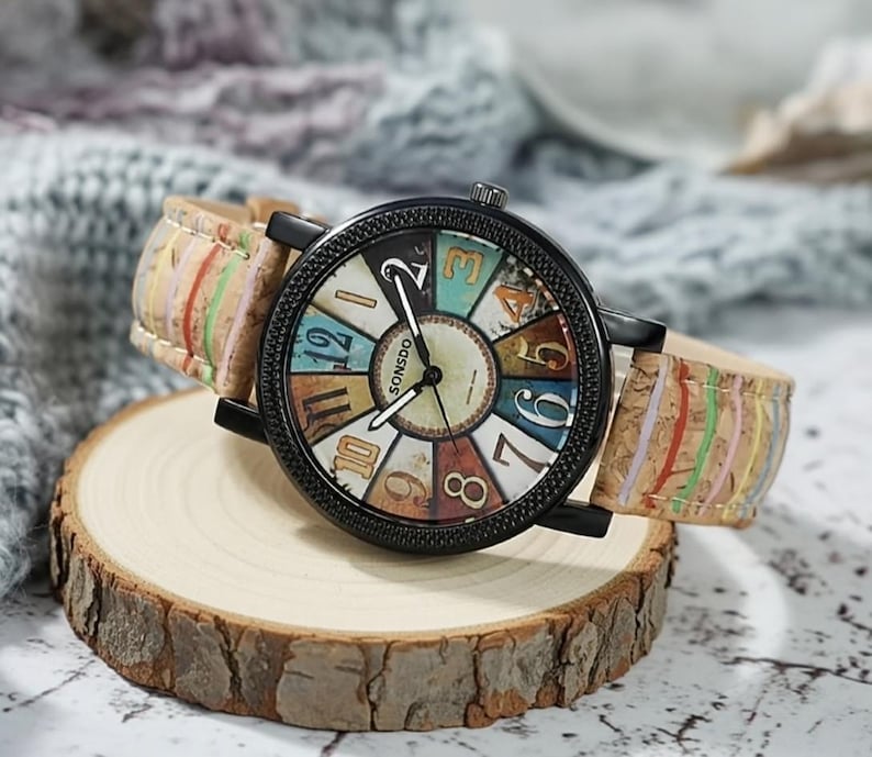 May include: A round wristwatch with a black case and a colorful face featuring the text "SONSDO". The watch has a cork band with colorful stripes and is displayed on a wooden slice.