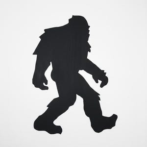 May include: A black silhouette of a Bigfoot figure, walking leftwards. The figure has a large head, broad shoulders, and thick legs, with a shaggy appearance. The background is plain white.