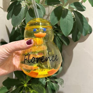 May include: A yellow duck-shaped tumbler with a clear straw. The tumbler features blue eyes and an orange beak. The name "Florence" is written in black script. The tumbler is held in front of green leafy plants.