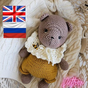 Peut inclure: Un ours en peluche crocheté, avec une tête et des membres marron clair, vêtu d'une tenue jaune et d'un col blanc à volants. Le col comporte une petite application d'abeille. L'ours est posé sur une couverture en tricot crème.