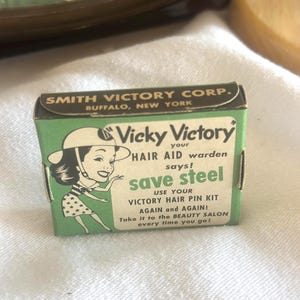 Op de afbeelding: Een vintage groene en witte doos voor "Vicky Victory" haarspeldkits. De doos toont een cartoonvrouw en de tekst "save steel" en "Take it to the BEAUTY SALON every time you go!"
