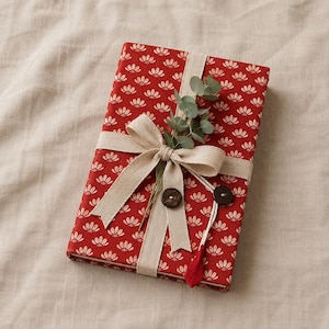 May include: A rectangular book wrapped in red fabric featuring a repeating white floral pattern. The book is tied with a beige ribbon bow, decorated with a small sprig of green eucalyptus leaves, two dark brown circular buttons, and a red fabric tag.
