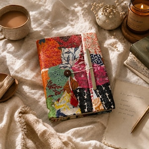 May include: A rectangular journal with a patchwork cover featuring various floral and geometric patterns in red, green, orange, pink, and black. The journal is secured with a string wrapped around a small brown button and a vertical metal clasp.