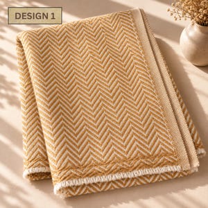 May include: A folded textile featuring a mustard yellow and cream white herringbone pattern. The fabric has a textured weave with a solid cream border along the edge and a fringed hem. The text "DESIGN 1" is printed in a box in the corner.