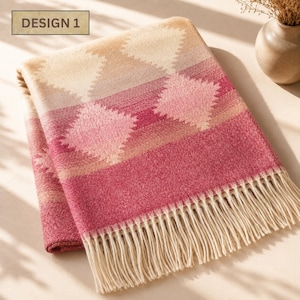 May include: A folded throw blanket featuring a geometric diamond pattern in shades of cream, beige, and deep pink. The blanket has a textured surface and is finished with long, cream-colored fringe along the bottom edge. The text DESIGN 1 is visible.