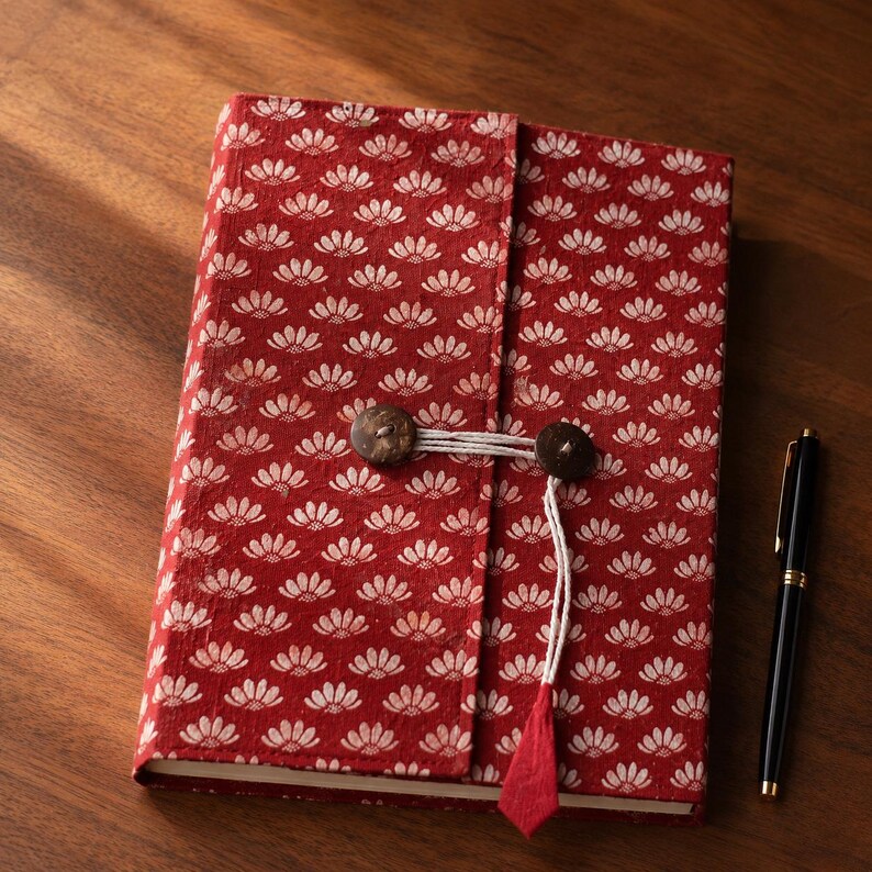 May include: A rectangular journal bound in red fabric with a repeating white floral pattern. The cover features a wrap-around closure secured by two dark brown circular buttons and a white cord. A black ballpoint pen lies next to the journal.