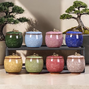 Può includere: Otto piccoli barattoli in ceramica con finitura smaltata a crepa in vari colori tra cui verde, blu, rosa, giallo e beige. Ogni barattolo presenta un corpo arrotondato, un coperchio abbinato con una piccola maniglia ad anello in metallo ottonato.