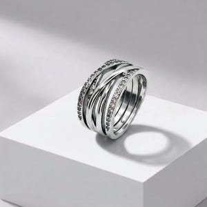 May include: A silver-toned multi-band ring featuring a crossover design with three distinct bands. Two of the bands are encrusted with small, clear, round-cut stones, while the central band is smooth and polished, creating a layered appearance.