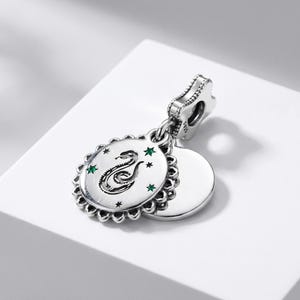 May include: A sterling silver charm featuring a circular pendant with a scalloped edge. The front displays an engraved snake motif surrounded by small green stones and star accents. A plain, smooth circular disc sits behind the main pendant.