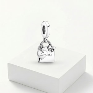 May include: A silver charm with a shopping bag design. The bag has a bow and the word "ORA" on it. A heart-shaped charm with the words "I love shopping" hangs from the bag. The charm is suspended from a silver loop.