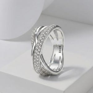 Könnte beinhalten: Ein silberfarbener Ring mit einem mehrbändigen Crossover-Design, bei dem ein Band mit kleinen klaren Steinen besetzt ist. Die Innenseite des Rings ist deutlich mit dem Text S925 geprägt, was die Metallzusammensetzung des Schmuckstücks angibt.
