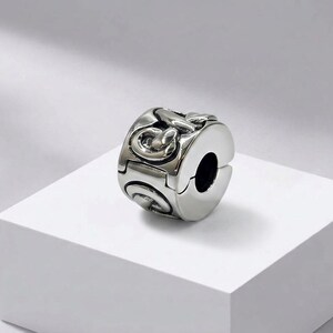 Könnte beinhalten: Ein silberfarbener Metall-Charm-Bead in zylindrischer Form mit einem aufklappbaren Scharniermechanismus. Die Außenfläche zeigt ein erhabenes, ineinandergreifendes Kettengliedmuster mit polierten Glanzlichtern und dunklen Vertiefungen.