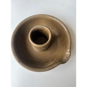 Pode incluir: Um castiçal de cerâmica marrom com base circular e copo cilíndrico central. O castiçal tem um pequeno bico na lateral. A peça tem uma aparência rústica e artesanal. Diâmetro: aprox. 15 cm.
