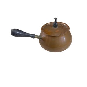 May include: A copper saucepan with a lid and a black handle. The small pot has a rounded body and a lid with a black knob. The handle is black and attached with a brass fitting. The saucepan is likely used for cooking.