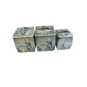 May include: Three vintage blue and white metal storage containers with lids. The containers feature windmill and sailboat scenes. The lids have handles. The containers are in varying sizes, with the largest measuring approximately 8 inches tall.