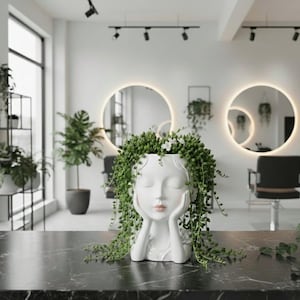 May include: A white ceramic planter shaped like a woman's face, with her hands supporting her cheeks. The planter is filled with cascading green plants and small white flowers. The planter is on a black marble surface.