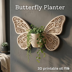 Op de afbeelding: Een beige vlindervormige wandplanter met ingewikkelde uitgesneden patronen. Een witte cilindrische pot bevat groene planten met hangende ranken. De tekst "Butterfly Planter" en "3D printable stl file" zijn zichtbaar.