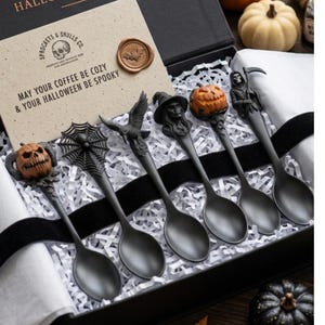 May include: A set of five matte black spoons with detailed Halloween-themed handles featuring a pumpkin, spider web, raven, witch, and grim reaper. The spoons are presented in a black gift box with a card that reads, May your coffee be cozy & your halloween be spooky.