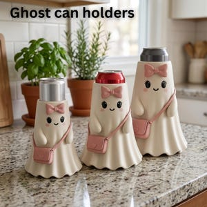 May include: Three off-white ghost-shaped can holders with the text Ghost can holders. Each ghost features a pink bow, a small pink crossbody bag, and a smiling face. The holders are designed to fit around standard beverage cans of varying sizes.