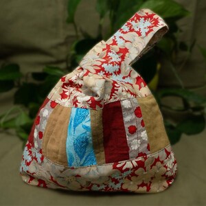 May include: A handmade patchwork bag with a handle. The bag features a variety of fabrics, including floral prints in red and cream, and solid and patterned stripes in brown, blue, and red. The bag is a unique, handcrafted accessory.