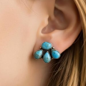 May include: A silver-toned stud earring featuring five turquoise stones. The stones are teardrop and oval shaped, arranged in a cluster. The earring is shown on an ear with blonde hair.