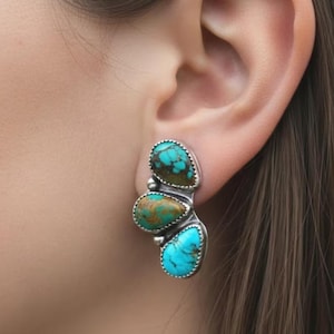 May include: A silver earring with three turquoise stones. The stones are teardrop-shaped and vary in shades of blue and green. The earring is worn on the ear.