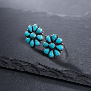 May include: A pair of silver stud earrings. Each earring has a flower design with turquoise-coloured, teardrop-shaped stones. The earrings are set against a dark, textured surface.