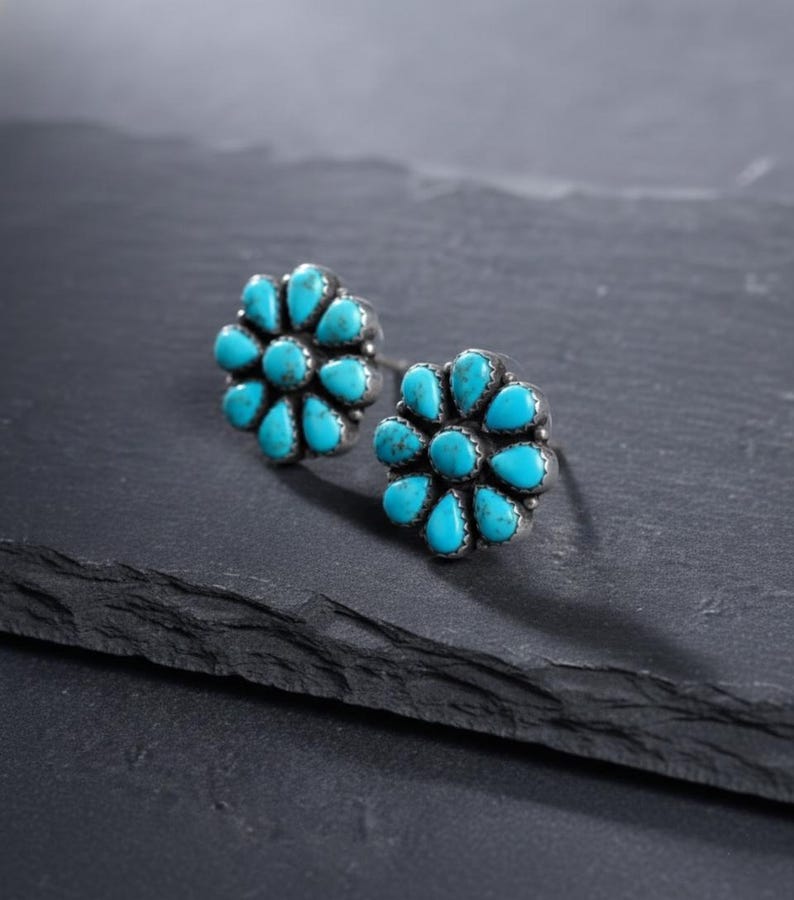May include: A pair of silver stud earrings. Each earring has a flower design with turquoise-coloured, teardrop-shaped stones. The earrings are set against a dark, textured surface.
