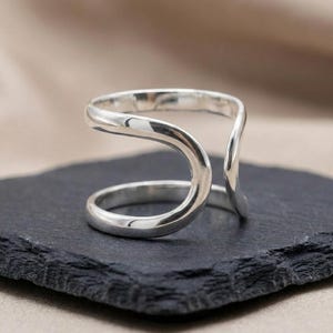 May include: A silver ring with a unique, open design. The ring features a curved, flowing shape that wraps around the finger, creating a modern and minimalist aesthetic. The ring is displayed on a dark, textured surface.