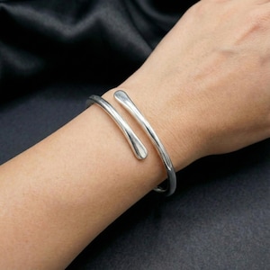 以下が含まれることがあります： オープンデザインのシルバーカラーのカフブレスレット。滑らかで光沢のある仕上げで手首に巻き付きます。ブレスレットの両端は丸みを帯びた水滴型をしています。