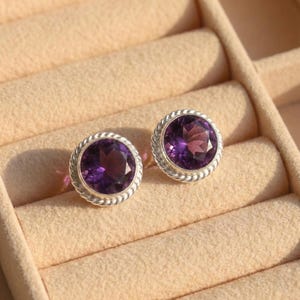May include: A pair of round amethyst stud earrings. Each earring features a deep purple gemstone set in a silver-colored, rope-textured bezel. The earrings are displayed in a beige jewelry box.